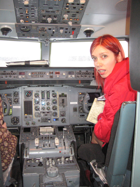 Kristīne sarkanā jakā sēž lidmašīnas pilotu kabīnē starp instrumentiem