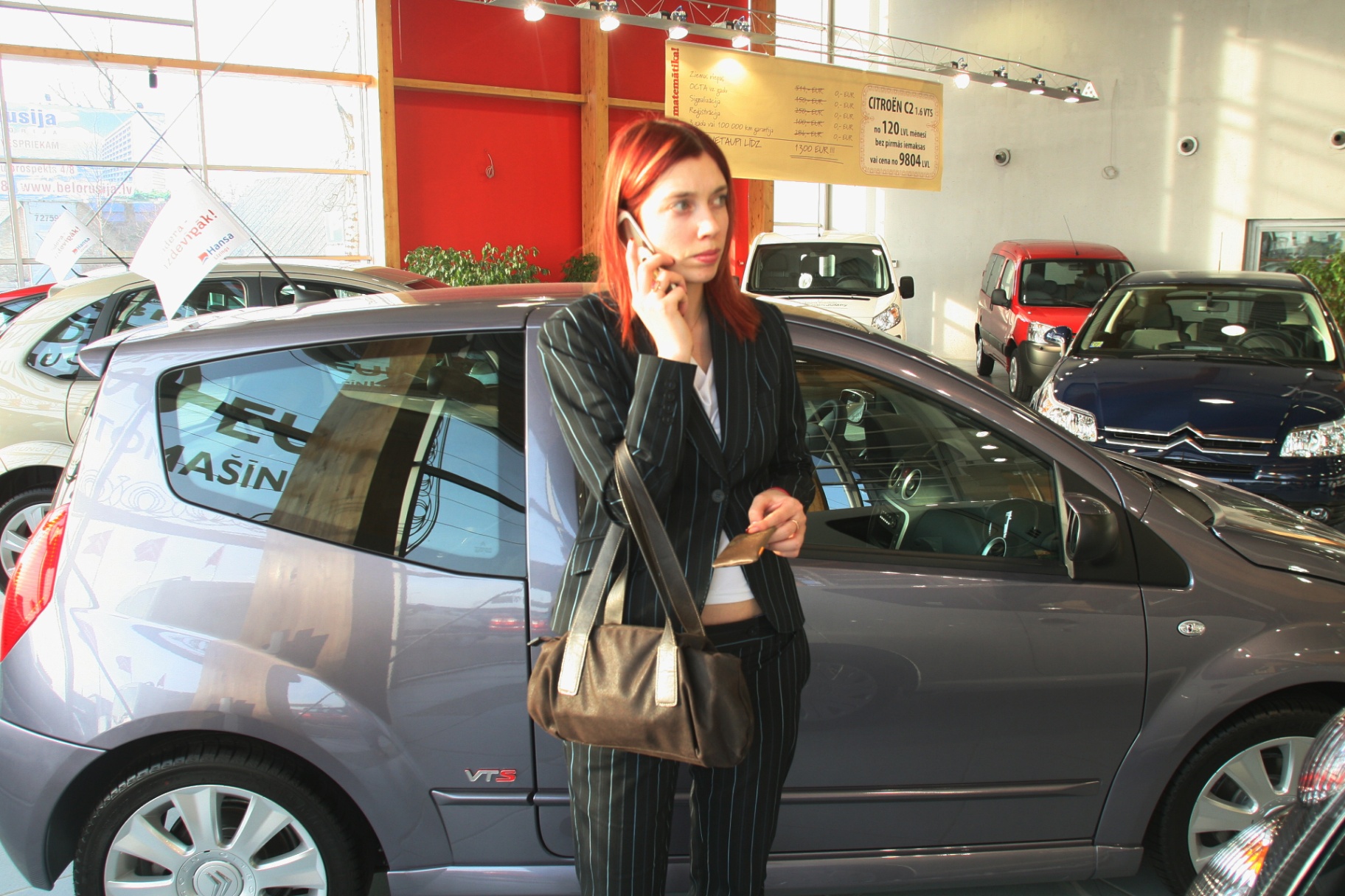 Kristīne biznesa uzvalkā ar mobilo telefonu pie auss stāv autosalonā pie automašīnas